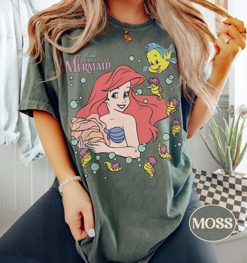 May include: Moss green t-shirt with a graphic of Ariel from Disney's The Little Mermaid. The design includes Ariel with red hair, a blue seashell bra, and Flounder. The shirt has the text "Disney The Little Mermaid".