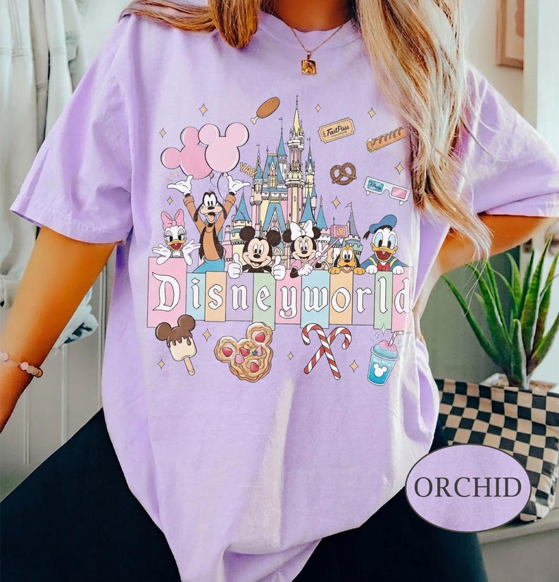 May include: Orchid-coloured t-shirt with a vibrant Disney World graphic. The design showcases cartoon characters such as Mickey and Minnie Mouse, Goofy, and Donald Duck, alongside a castle and food icons. The word "Disneyworld" is clearly visible.