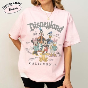 May include: A pale pink t-shirt with a Disneyland graphic featuring Mickey Mouse, Minnie Mouse, Donald Duck, Daisy Duck, Goofy, and Pluto. The shirt includes the text "Disneyland California" and "EST. 1955". Made of Comfort Colors material.