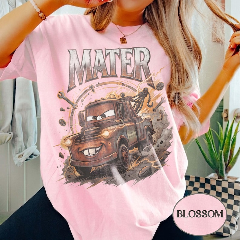 May include: A light pink t-shirt featuring a graphic print of the character Mater from the movie Cars. The design includes the text MATER in a metallic font above the brown tow truck, surrounded by tools, gears, and dust clouds on the shirt front.