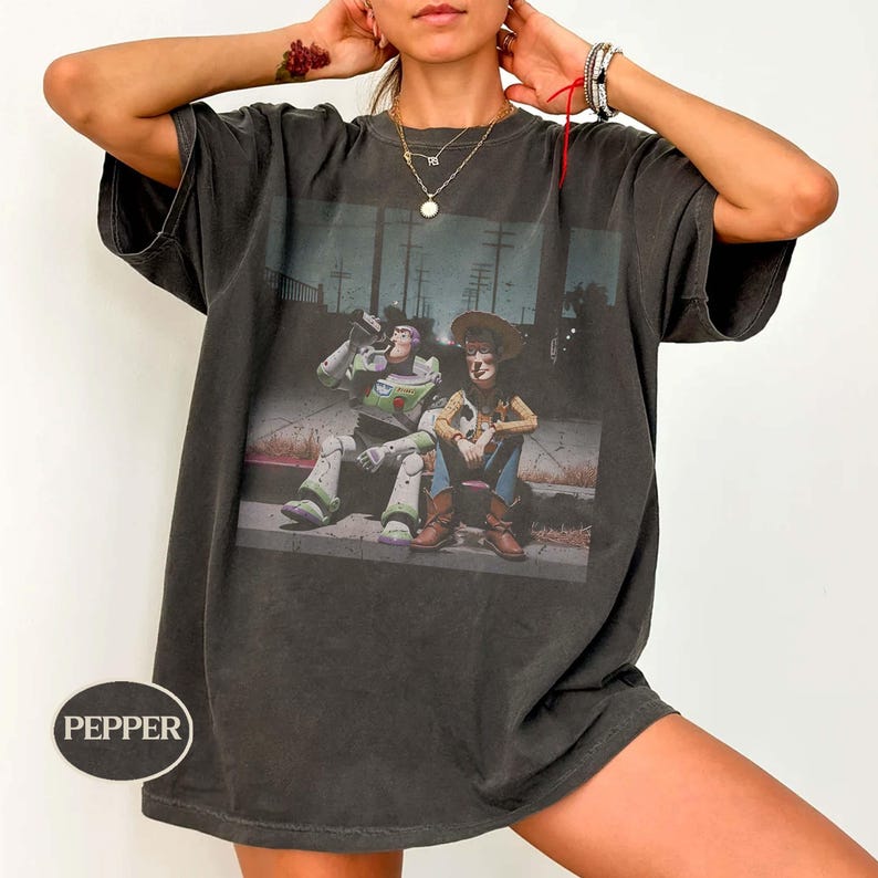 May include: A charcoal gray short-sleeve t-shirt featuring a graphic print of the characters Buzz Lightyear and Woody sitting together in a dark, outdoor setting. The shirt has a crew neckline and a small oval logo with the text PEPPER.