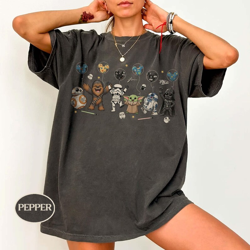 May include: Dark grey t-shirt featuring a graphic of Star Wars characters holding balloons. The characters include BB-8, Chewbacca, a Stormtrooper, Grogu, R2-D2, and Darth Vader. The word "Pepper" is printed on the bottom left of the shirt.