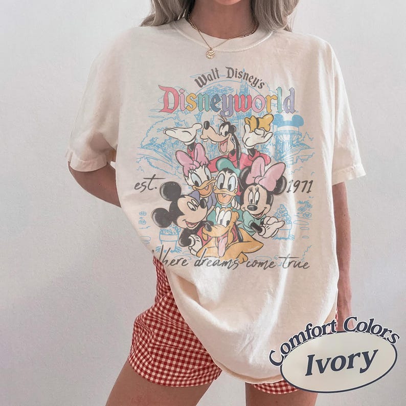 May include: Ivory-colored t-shirt featuring a colorful graphic of Disney characters, including Mickey Mouse, Minnie Mouse, and Donald Duck. The shirt has the text "Walt Disney's Disneyworld" and "est. 1971" printed on it. The phrase "Where dreams come true" is also printed on the shirt.