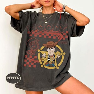 May include: A charcoal gray short-sleeve t-shirt featuring a graphic print of the character Woody from Toy Story. The design includes a red checkered pattern, the text Woody 1995, and a yellow sheriff star emblem. The shirt has a crew neckline.