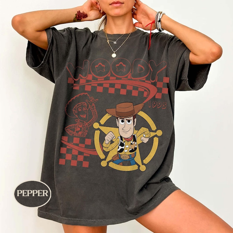 May include: A charcoal gray short-sleeve t-shirt featuring a graphic print of the character Woody from Toy Story. The design includes a red checkered pattern, the text Woody 1995, and a yellow sheriff star emblem. The shirt has a crew neckline.