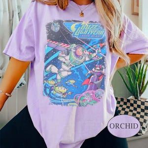Puede incluir: Camiseta morada orqu&iacute;dea con un gr&aacute;fico de Buzz Lightyear de la pel&iacute;cula Toy Story. El gr&aacute;fico incluye el texto "Buzz Lightyear Laser Blast" e im&aacute;genes de los personajes. La camiseta es de manga corta y cuello redondo.