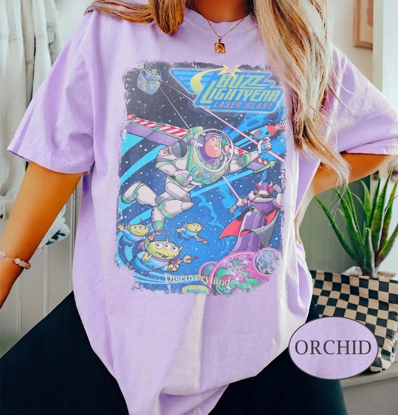 Puede incluir: Camiseta morada orqu&iacute;dea con un gr&aacute;fico de Buzz Lightyear de la pel&iacute;cula Toy Story. El gr&aacute;fico incluye el texto "Buzz Lightyear Laser Blast" e im&aacute;genes de los personajes. La camiseta es de manga corta y cuello redondo.