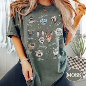May include: A dark green t-shirt featuring a graphic print of various animated animal characters from Zootopia, including Judy Hopps, Nick Wilde, and Flash, with their names written in script text alongside small star and accessory icons.
