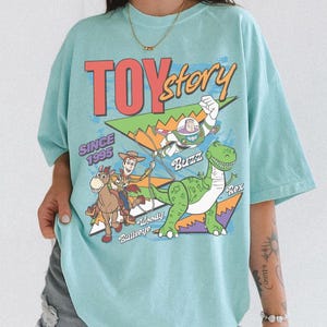 May include: A light blue t-shirt featuring a graphic from the movie Toy Story. The graphic includes the words "TOY STORY" and "SINCE 1995". The graphic also features the characters Buzz Lightyear, Woody, Bullseye, and Rex.