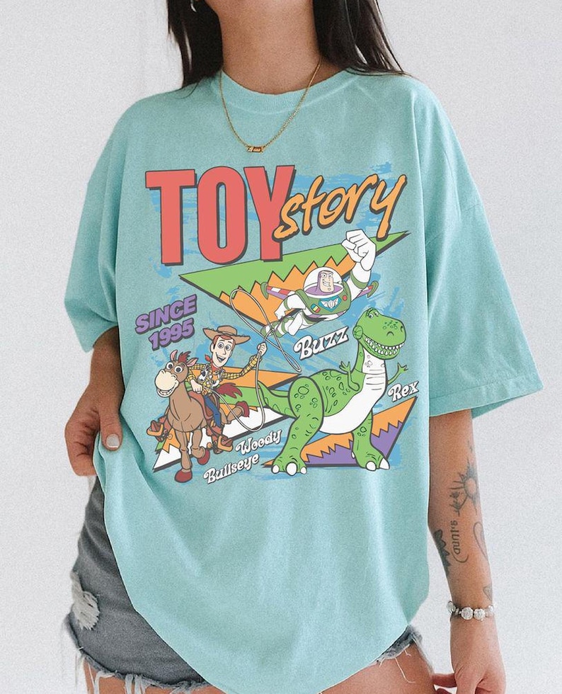 May include: A light blue t-shirt featuring a graphic from the movie Toy Story. The graphic includes the words "TOY STORY" and "SINCE 1995". The graphic also features the characters Buzz Lightyear, Woody, Bullseye, and Rex.