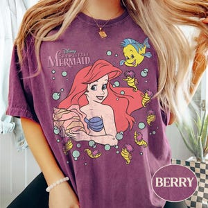 May include: Berry-coloured t-shirt featuring a graphic of Ariel from Disney's The Little Mermaid. The design includes Ariel with red hair, a blue seashell bra, and Flounder. The shirt has a vintage, washed look.