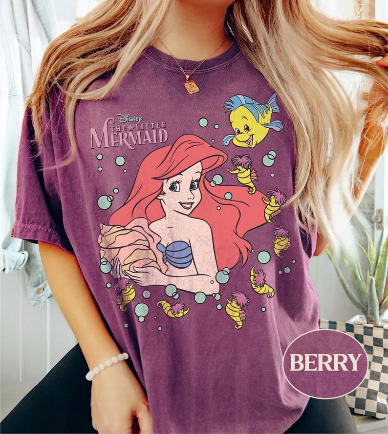 May include: Berry-coloured t-shirt featuring a graphic of Ariel from Disney's The Little Mermaid. The design includes Ariel with red hair, a blue seashell bra, and Flounder. The shirt has a vintage, washed look.