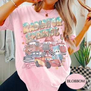 K&ouml;nnte beinhalten: Hellrosa T-Shirt mit einem Aufdruck von Charakteren aus dem Film Cars, darunter Lightning McQueen. Der Text "Radiator Springs" ist &uuml;ber den Charakteren abgebildet. Das Shirt hat auch den Text "Lightning McQueen" unten.