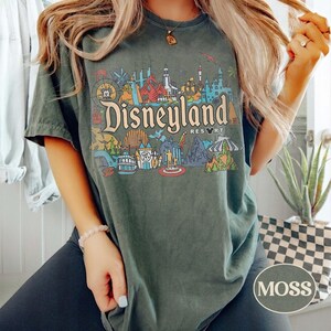 Op de afbeelding: Donkergroen T-shirt met een kleurrijke Disneyland Resort-afbeelding. Het ontwerp bevat verschillende parkattracties en het Disneyland-logo. Het shirt heeft een relaxte pasvorm en korte mouwen. Het woord "MOSS" staat rechtsonder opgedrukt.