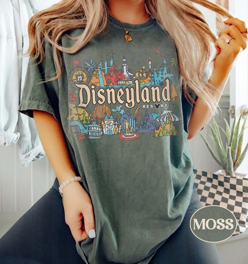 Op de afbeelding: Donkergroen T-shirt met een kleurrijke Disneyland Resort-afbeelding. Het ontwerp bevat verschillende parkattracties en het Disneyland-logo. Het shirt heeft een relaxte pasvorm en korte mouwen. Het woord "MOSS" staat rechtsonder opgedrukt.