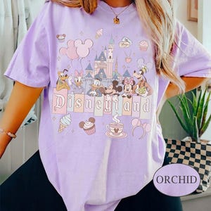 Może przedstawiać: Fioletowa koszulka z nadrukiem Disneyland. Projekt zawiera postacie z kresk&oacute;wek, zamek i napis "Disneyland". Dodatkowe grafiki obejmują rożki lod&oacute;w, babeczki i uszy Myszki Miki. Koszulka jest kr&oacute;tka i ma okrągły dekolt.