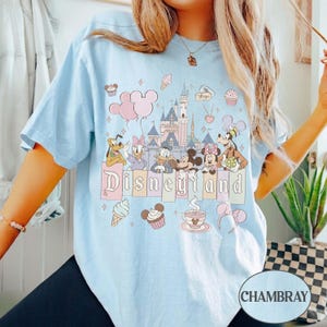 Może przedstawiać: Jasnoniebieska koszulka z grafiką Disneyland. Projekt zawiera postacie Disneya, takie jak Myszka Miki i Myszka Minnie, oraz zamek. Słowo "Disneyland" jest napisane pastelową czcionką, z babeczkami, lodami i balonami.