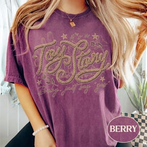 May include: A berry-colored short-sleeve t-shirt featuring a graphic design with the text "it's a Toy Story" in a rope-like font, accompanied by the phrase "Baby, just say yes" and illustrations of cacti, stars, and small flowers.
