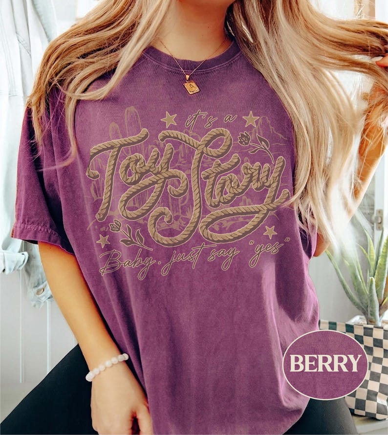 May include: A berry-colored short-sleeve t-shirt featuring a graphic design with the text "it's a Toy Story" in a rope-like font, accompanied by the phrase "Baby, just say yes" and illustrations of cacti, stars, and small flowers.