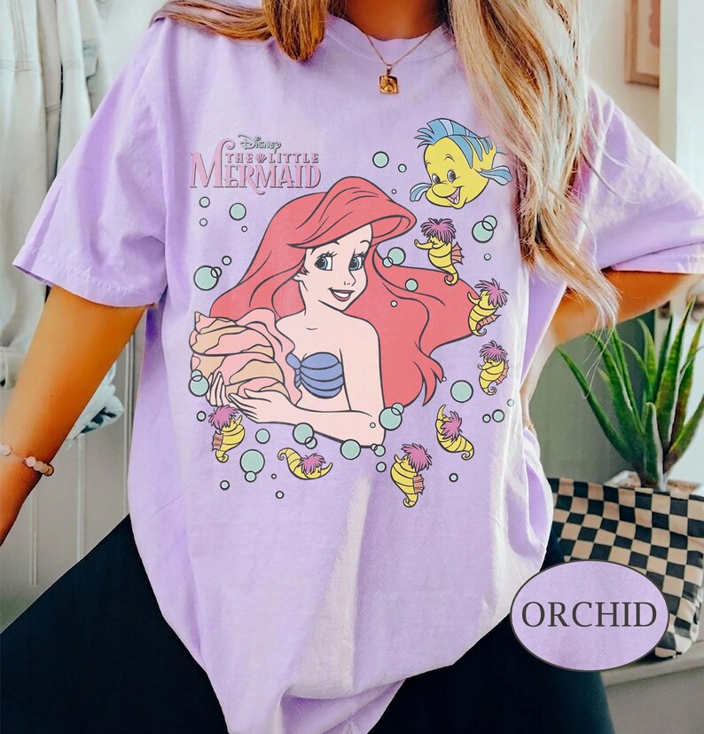 May include: Lilac t-shirt with a graphic of Ariel from Disney's The Little Mermaid. The design includes Ariel, Flounder, and seahorse characters. The shirt has a relaxed fit and short sleeves.