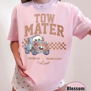 May include: A light pink t-shirt featuring a graphic of the character Tow Mater from the movie Cars. The text reads TOW MATER, PISTON CUP RACING SERIES, and 5 TIME CHAMP, accompanied by a gold trophy icon and a brown checkered pattern design.