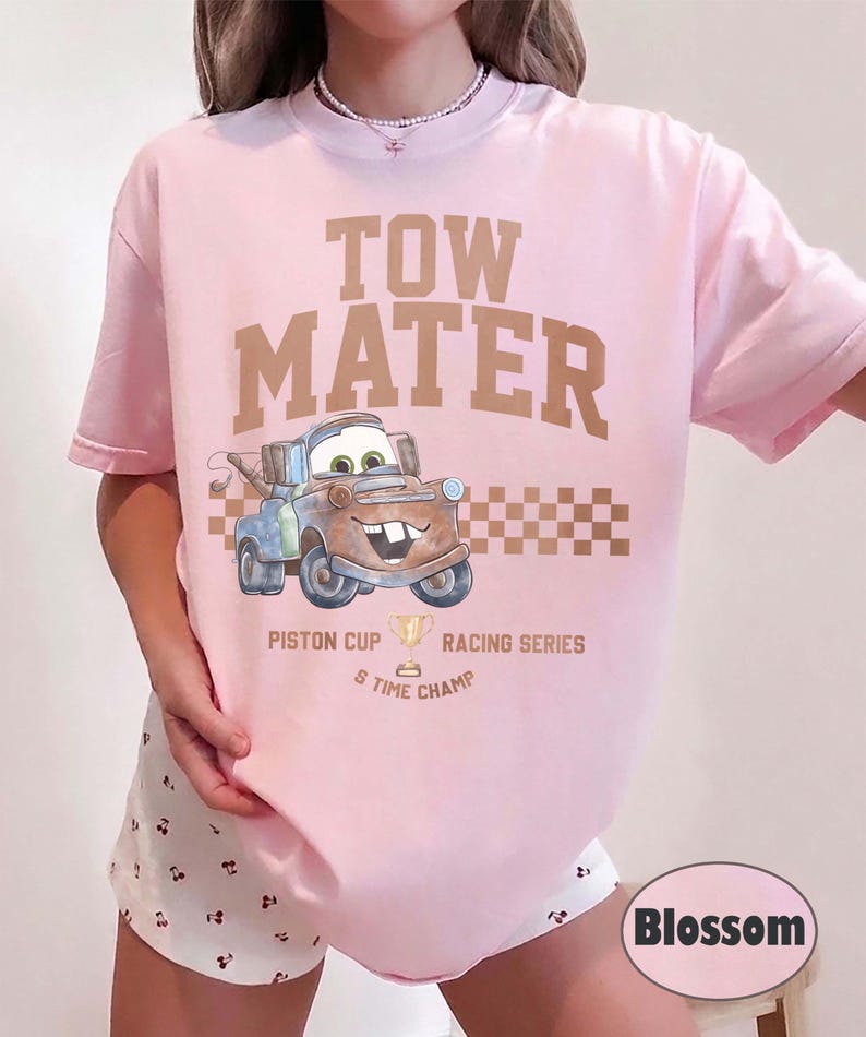 May include: A light pink t-shirt featuring a graphic of the character Tow Mater from the movie Cars. The text reads TOW MATER, PISTON CUP RACING SERIES, and 5 TIME CHAMP, accompanied by a gold trophy icon and a brown checkered pattern design.