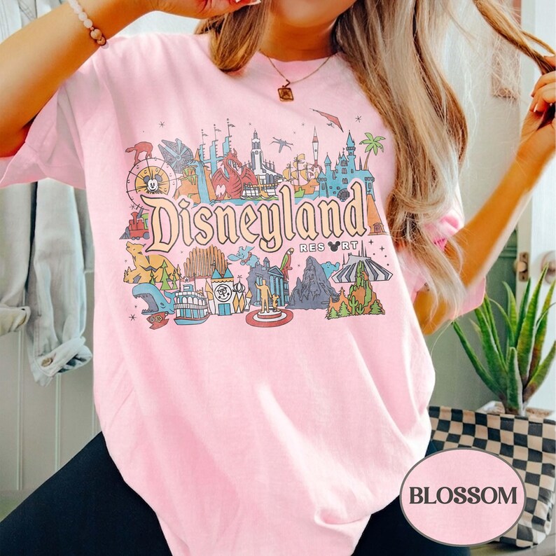 Op de afbeelding: Lichtroze T-shirt met een kleurrijke Disneyland-afbeelding. De afbeelding bevat het Disneyland Resort-logo, iconische bezienswaardigheden en stripfiguren. Het shirt is een comfortabele, casual stijl.