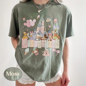 Może przedstawiać: Zielona koszulka w kolorze mchu z kolorową grafiką Disneyland. Projekt zawiera postacie z kresk&oacute;wek, zamek i napis "Disneyland" w pastelowych kolorach. Dodatkowa grafika obejmuje rożki lod&oacute;w, babeczki i filiżankę herbaty.