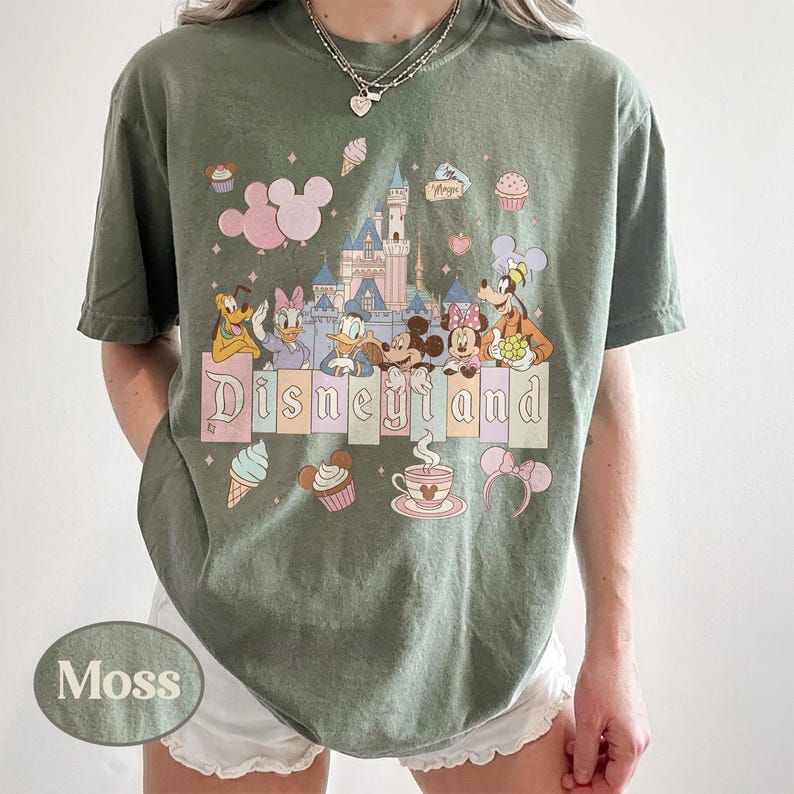 Może przedstawiać: Zielona koszulka w kolorze mchu z kolorową grafiką Disneyland. Projekt zawiera postacie z kresk&oacute;wek, zamek i napis "Disneyland" w pastelowych kolorach. Dodatkowa grafika obejmuje rożki lod&oacute;w, babeczki i filiżankę herbaty.