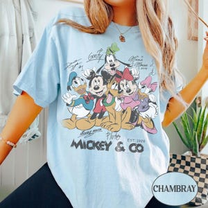 K&ouml;nnte beinhalten: Hellblaues T-Shirt mit einer Grafik von Micky Maus und ihren Freunden, darunter Donald Duck, Goofy, Minnie Maus und Daisy Duck. Das Design enth&auml;lt Unterschriften und den Text "Mickey & Co. EST. 1928".