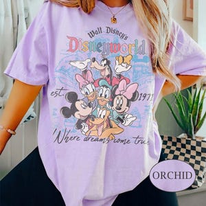 May include: Orchid-colored t-shirt featuring a graphic of Disney characters, including Mickey Mouse, Minnie Mouse, Donald Duck, and Goofy. The design includes the text "Walt Disney's Disneyworld" and "Where dreams come true."