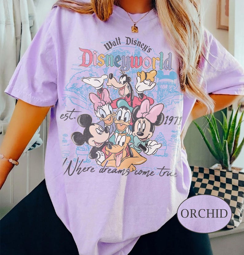 May include: Orchid-colored t-shirt featuring a graphic of Disney characters, including Mickey Mouse, Minnie Mouse, Donald Duck, and Goofy. The design includes the text "Walt Disney's Disneyworld" and "Where dreams come true."