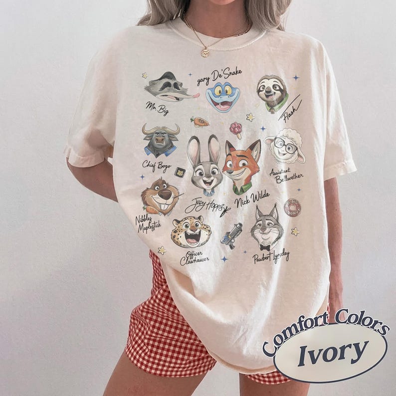 May include: An ivory short-sleeve t-shirt featuring a graphic print of various animated animal characters including a rabbit, fox, sloth, and sheep. The text includes names like Judy Hopps, Nick Wilde, Mr. Big, and Flash, accompanied by small star icons.