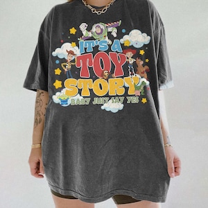Peut inclure: Un t-shirt gris anthracite &agrave; manches courtes avec une impression graphique color&eacute;e de personnages de Toy Story, dont Woody, Buzz l'&Eacute;clair et Jessie. Le texte indique IT'S A TOY STORY BABY JUST SAY YES, entour&eacute; de nuages et d'&eacute;toiles jaunes.