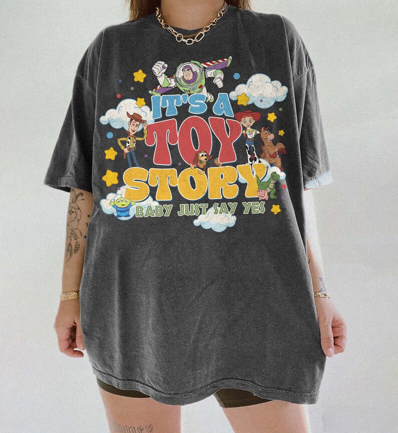 Peut inclure: Un t-shirt gris anthracite &agrave; manches courtes avec une impression graphique color&eacute;e de personnages de Toy Story, dont Woody, Buzz l'&Eacute;clair et Jessie. Le texte indique IT'S A TOY STORY BABY JUST SAY YES, entour&eacute; de nuages et d'&eacute;toiles jaunes.