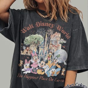 May include: Dark gray t-shirt featuring a colorful graphic of the Walt Disney World castle, characters, and fireworks. The text "Walt Disney World" and "The Happiest Place On Earth" are printed on the front. The shirt is labeled "Comfort Colors Pepper".