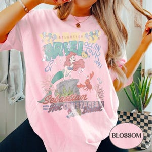 May include: A light pink t-shirt featuring a graphic print of Ariel, Flounder, and Sebastian from The Little Mermaid. The text reads Atlantica Ocean Tour 1991, Sebastian with the Hot Crustacean Band. The design includes bubbles and seaweed.