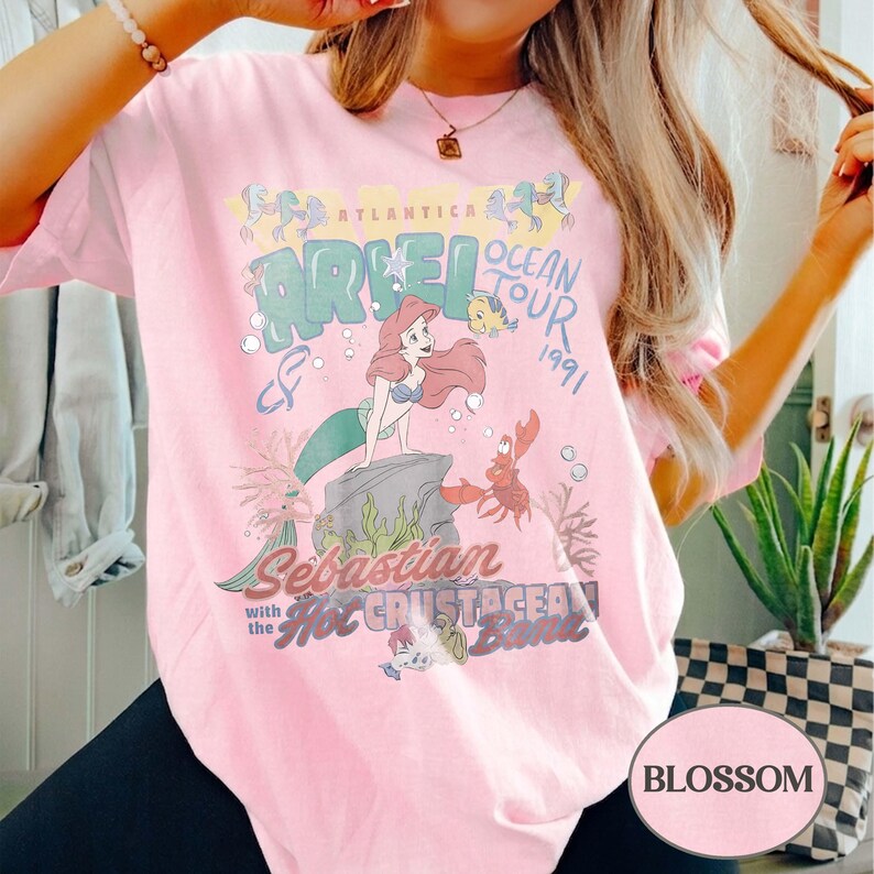 May include: A light pink t-shirt featuring a graphic print of Ariel, Flounder, and Sebastian from The Little Mermaid. The text reads Atlantica Ocean Tour 1991, Sebastian with the Hot Crustacean Band. The design includes bubbles and seaweed.
