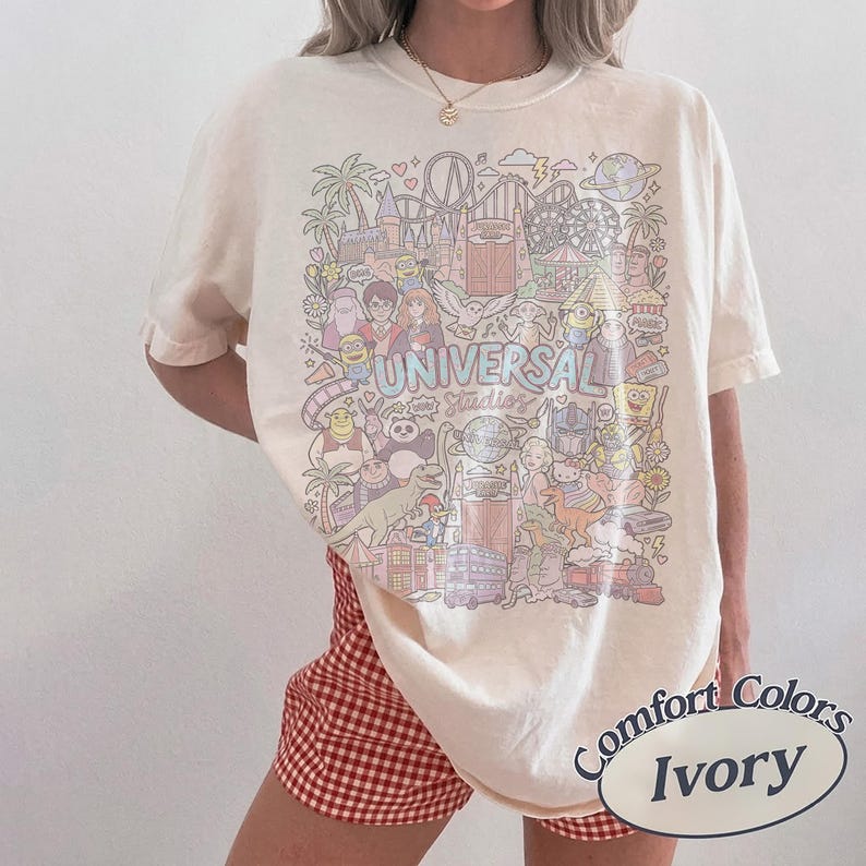 K&ouml;nnte beinhalten: Elfenbeinfarbenes T-Shirt mit einer farbenfrohen Grafik von Universal Studios-Charakteren und -Attraktionen. Das Shirt tr&auml;gt den Text "UNIVERSAL Studios" in einer Pastellschrift. Das Shirt ist von der Marke Comfort Colors.