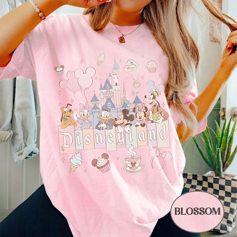 Może przedstawiać: Jasnor&oacute;żowa koszulka z grafiką Disneyland. Projekt zawiera postacie z kresk&oacute;wek, zamek i napis "Disneyland" w fantazyjnej czcionce. Dodatkowe elementy to balony Myszki Miki, rożki lod&oacute;w i babeczki.