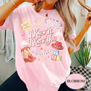 May include: A light pink t-shirt featuring a graphic print with the text I KNEW IT I KNEW YOU, a yellow sheriff star with INFINITY AND BEYOND, a beaded bracelet with YOU'VE GOT A FRIEND IN ME, red cowboy boots, a red hat, and alphabet blocks.