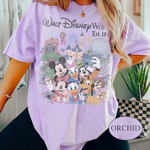 May include: A light purple t-shirt featuring a graphic print of Walt Disney World Est. 1971 with various cartoon characters including Mickey Mouse, Minnie Mouse, Donald Duck, Daisy Duck, Goofy, Pluto, and Chip and Dale in front of park landmarks.