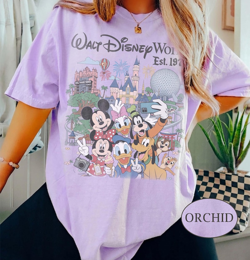 May include: A light purple t-shirt featuring a graphic print of Walt Disney World Est. 1971 with various cartoon characters including Mickey Mouse, Minnie Mouse, Donald Duck, Daisy Duck, Goofy, Pluto, and Chip and Dale in front of park landmarks.