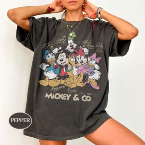 K&ouml;nnte beinhalten: Dunkelgraues T-Shirt mit einer Grafik von Micky Maus und ihren Freunden, darunter Donald Duck, Goofy, Minnie Maus und Pluto. Das Shirt tr&auml;gt den Text "Mickey & Co. Est. 1928" und Unterschriften der Charaktere. Das Shirt ist &uuml;bergro&szlig;.
