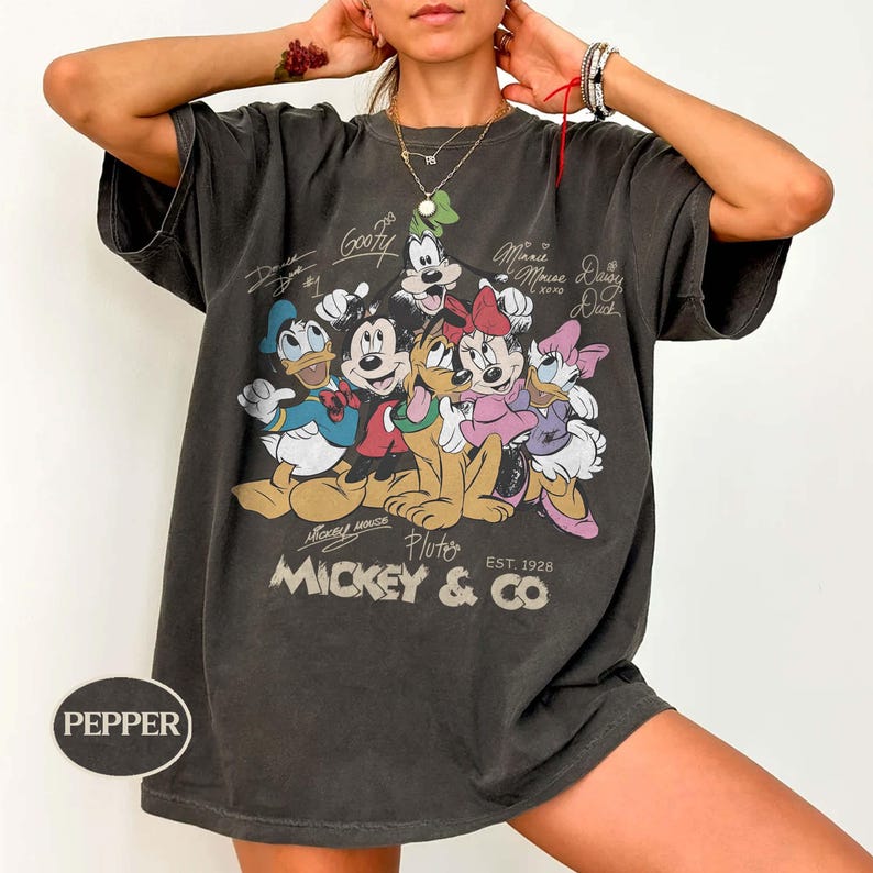 K&ouml;nnte beinhalten: Dunkelgraues T-Shirt mit einer Grafik von Micky Maus und ihren Freunden, darunter Donald Duck, Goofy, Minnie Maus und Pluto. Das Shirt tr&auml;gt den Text "Mickey & Co. Est. 1928" und Unterschriften der Charaktere. Das Shirt ist &uuml;bergro&szlig;.
