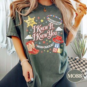 May include: A dark green t-shirt featuring the text I KNEW IT, I KNEW YOU, TOY STORY, INFINITY AND BEYOND, and YOU'VE GOT A FRIEND IN ME. The graphic includes a yellow star, a beaded bracelet, a red cowboy hat, red cowboy boots, and alphabet blocks.