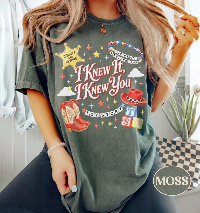May include: A dark green t-shirt featuring the text I KNEW IT, I KNEW YOU, TOY STORY, INFINITY AND BEYOND, and YOU'VE GOT A FRIEND IN ME. The graphic includes a yellow star, a beaded bracelet, a red cowboy hat, red cowboy boots, and alphabet blocks.