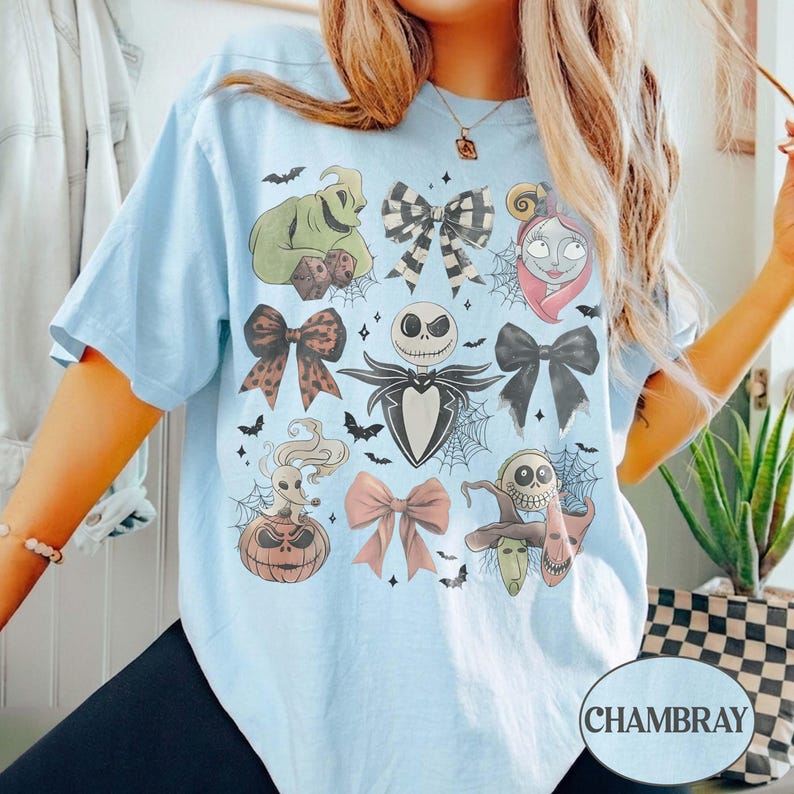 May include: A light blue t-shirt featuring a graphic print of characters and bows from The Nightmare Before Christmas. The design includes Jack Skellington, Sally, Oogie Boogie, Zero, and various bows, all surrounded by small bats and spiderwebs.