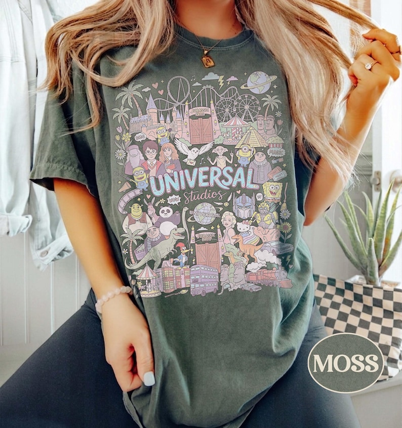 K&ouml;nnte beinhalten: Moosgr&uuml;nes T-Shirt mit einer farbenfrohen Grafik von Universal Studios-Charakteren und -Attraktionen. Das Design enth&auml;lt den Text "UNIVERSAL Studios" und verschiedene Cartoonfiguren. Das Shirt hat eine lockere Passform.
