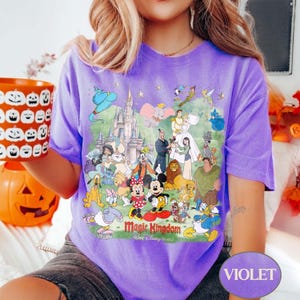 May include: A violet t-shirt featuring a colorful graphic of Disney characters and the text "Magic Kingdom". The design includes Mickey and Minnie Mouse, Donald Duck, and other beloved characters. The shirt is a comfortable, casual wear.